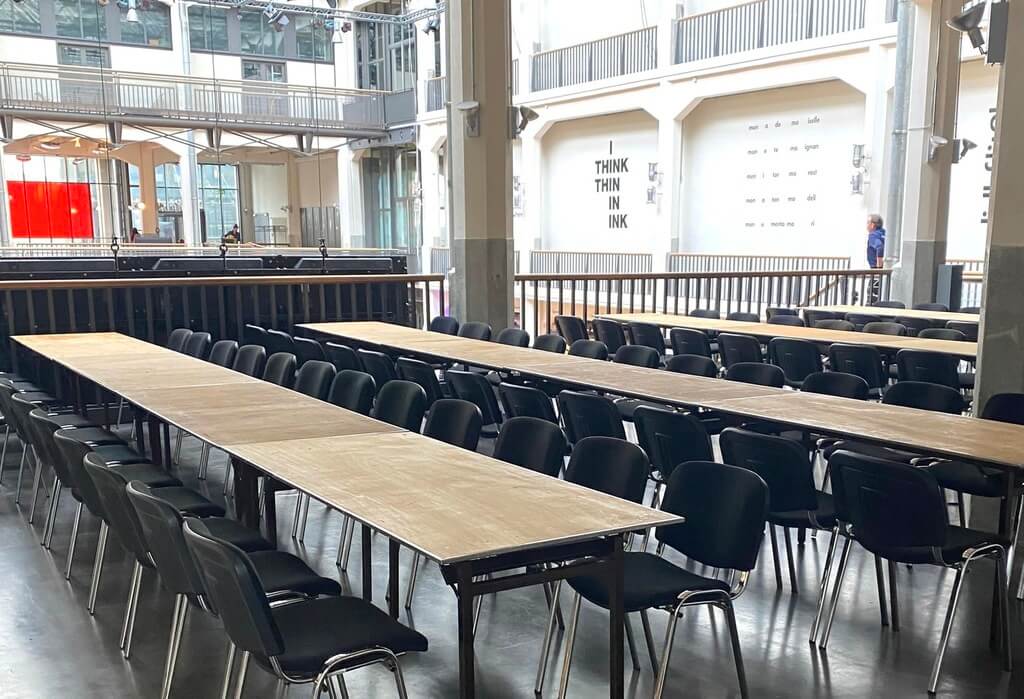 In einer hellen, modernen Halle sind Sitztische und schwarze Stühle in Reihen für Veranstaltungen aufgestellt