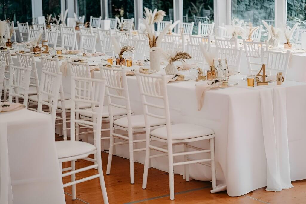 Elegante weiße Tafel mit weißen Stühlen, neutralen Mitteldekorationen und goldenen Akzenten, festlich gedeckt für eine Hochzeit oder ein Event