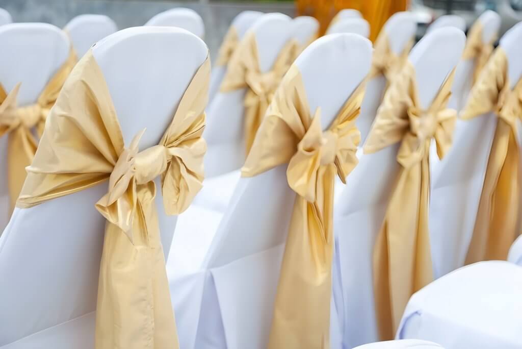 Weiße Stühle in Reihen mit großen goldenen Schleifen und eleganten Stuhlhussen Deluxe, ideal für festliche Anlässe oder Stuhlschleifen mieten